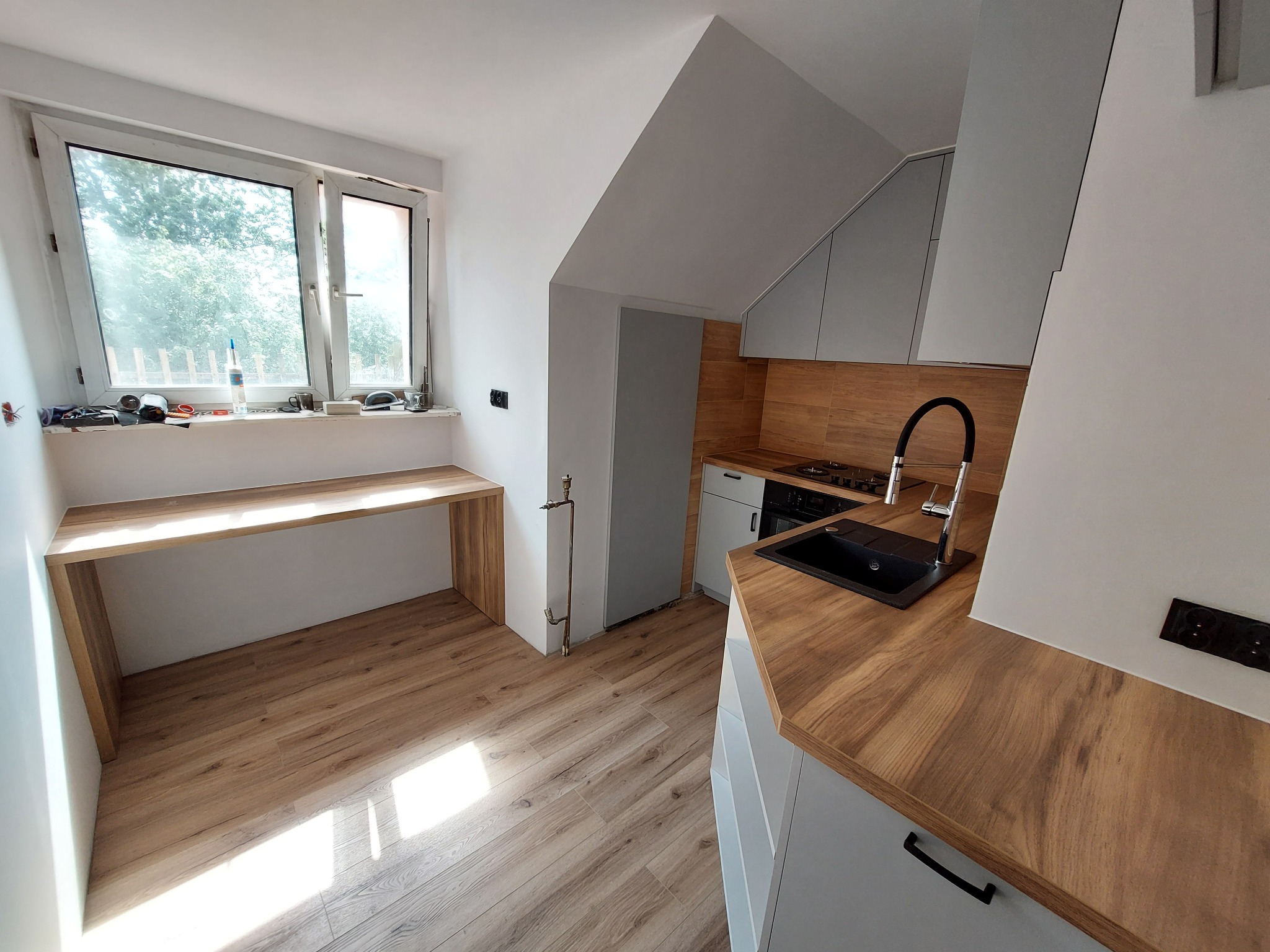 Jasna kuchnia w skosach z drewnianym blatem i podłogą. Okno z blatem roboczym. Szare szafki i czarny zlew. Ujęcie z perspektywy podłogi, widok na całe pomieszczenie.