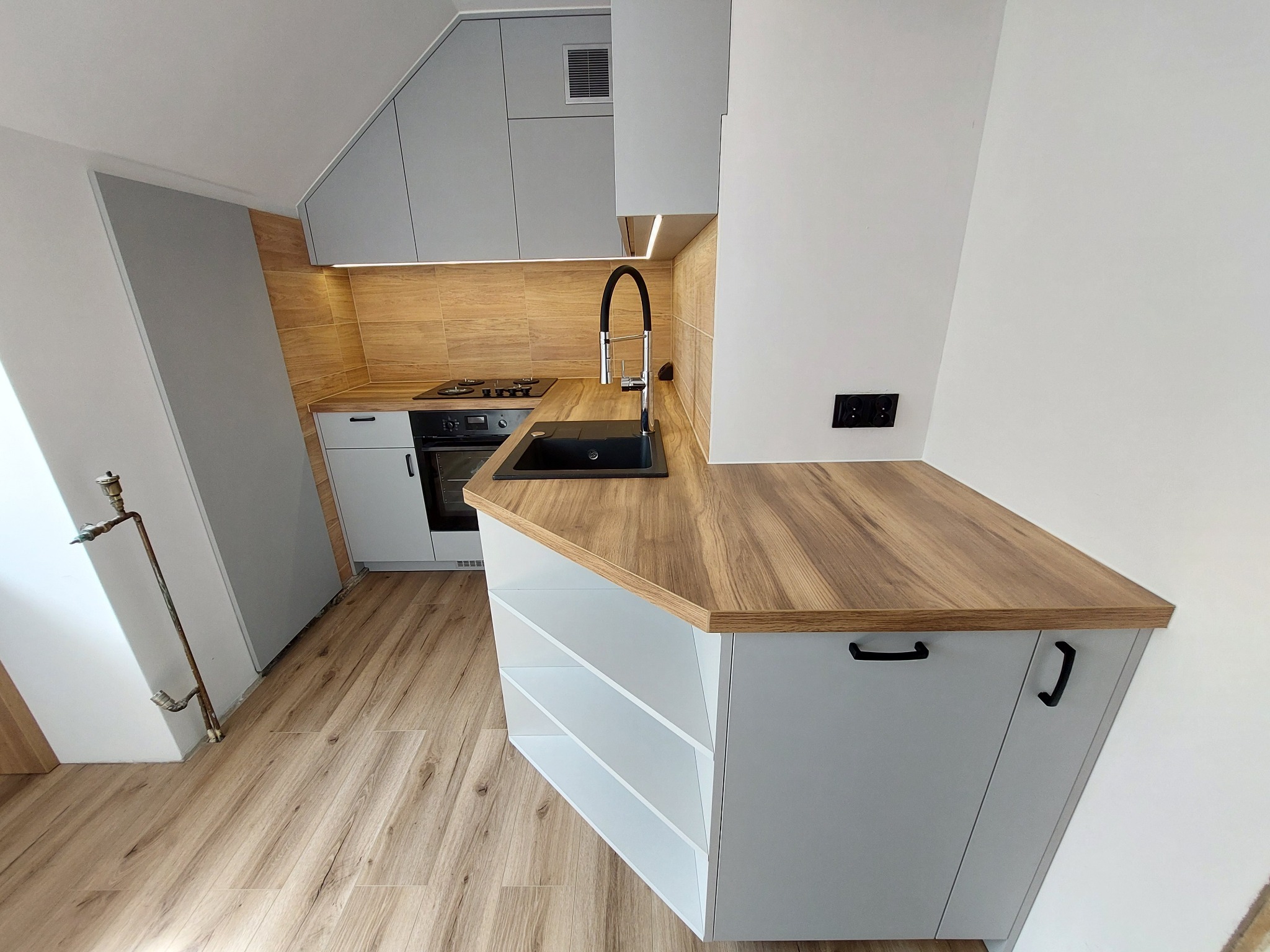 Szara kuchnia z drewnianym blatem i płytkami imitującymi drewno na ścianie. Uwagę przykuwa czarny zlew i bateria. Widoczna płyta grzewcza i piekarnik.