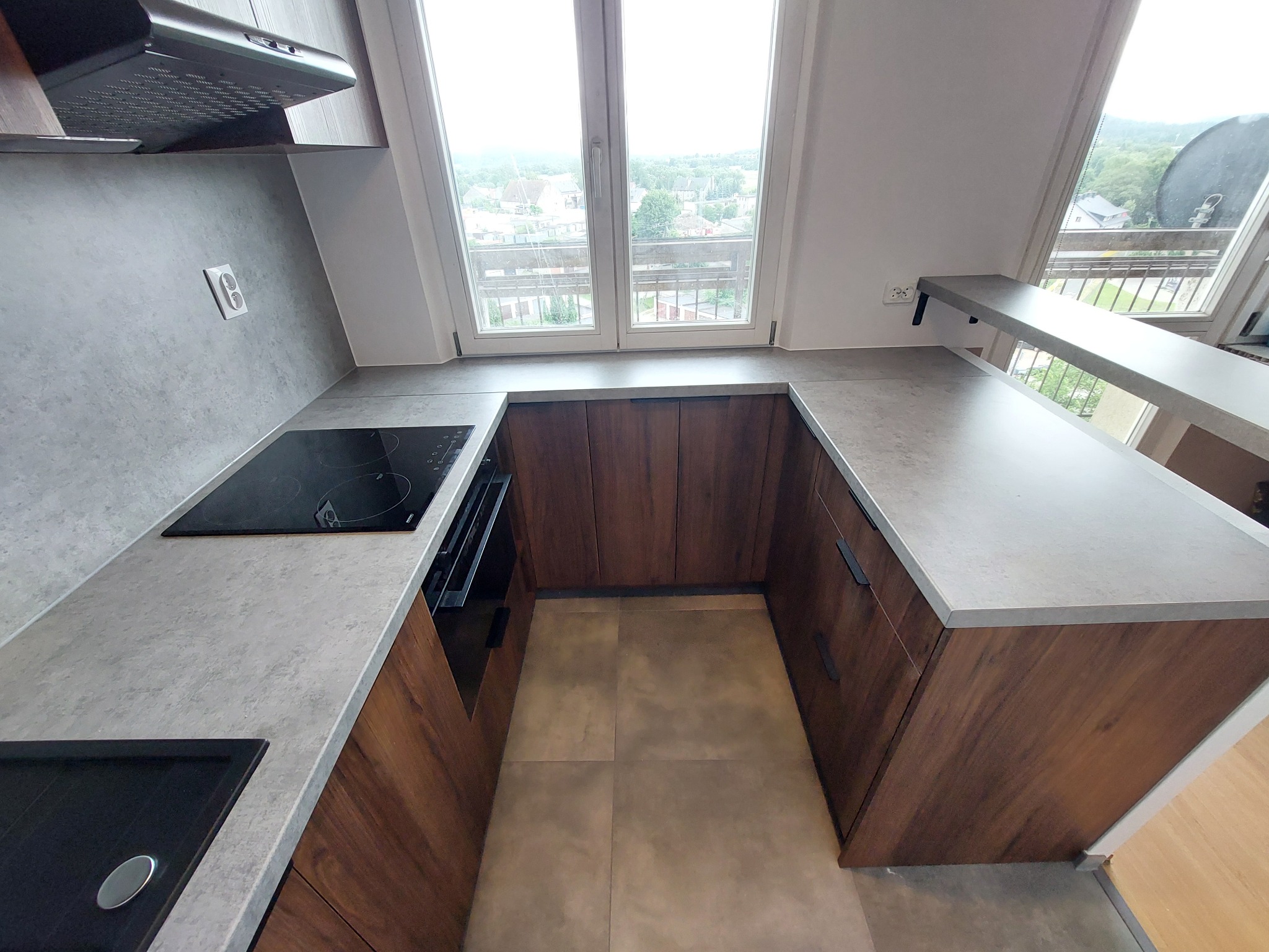 Nowoczesna kuchnia z drewnianymi szafkami i szarym blatem, płyta indukcyjna i zlewozmywak wkomponowane w blat. Widok na okno z zabudową kuchenną pod nim.