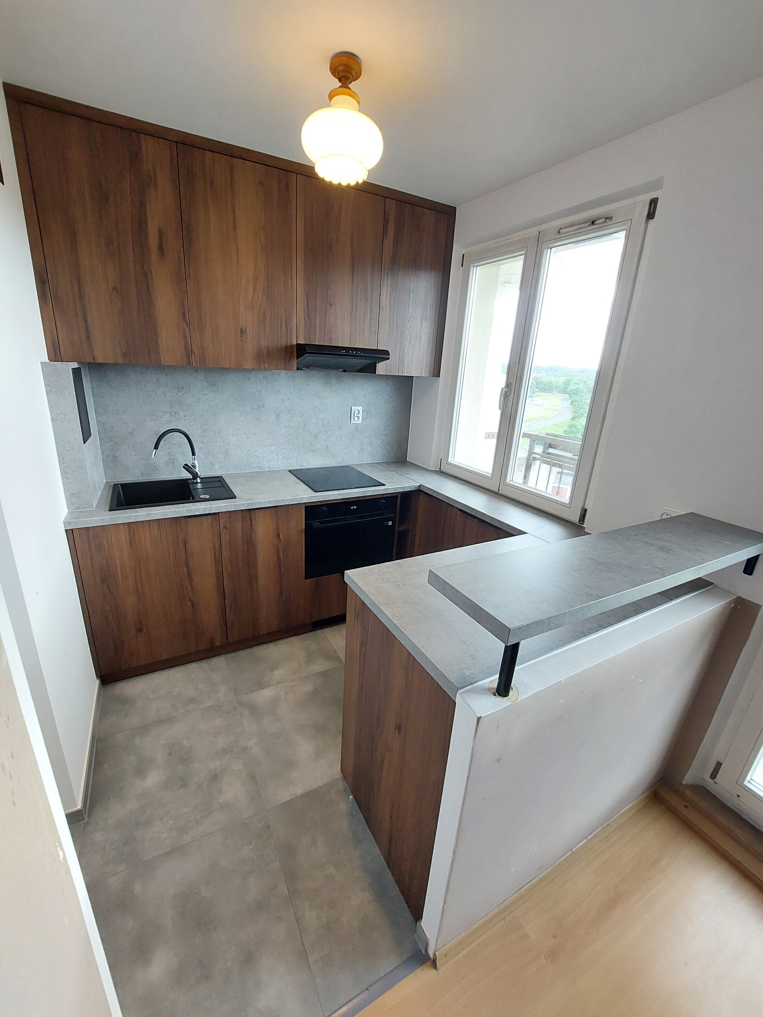 Nowoczesna kuchnia z drewnianymi szafkami i szarym blatem, czarny zlew i płyta indukcyjna, oświetlenie sufitowe, widok na zewnątrz przez okno. Ustawienie w kształcie litery U.