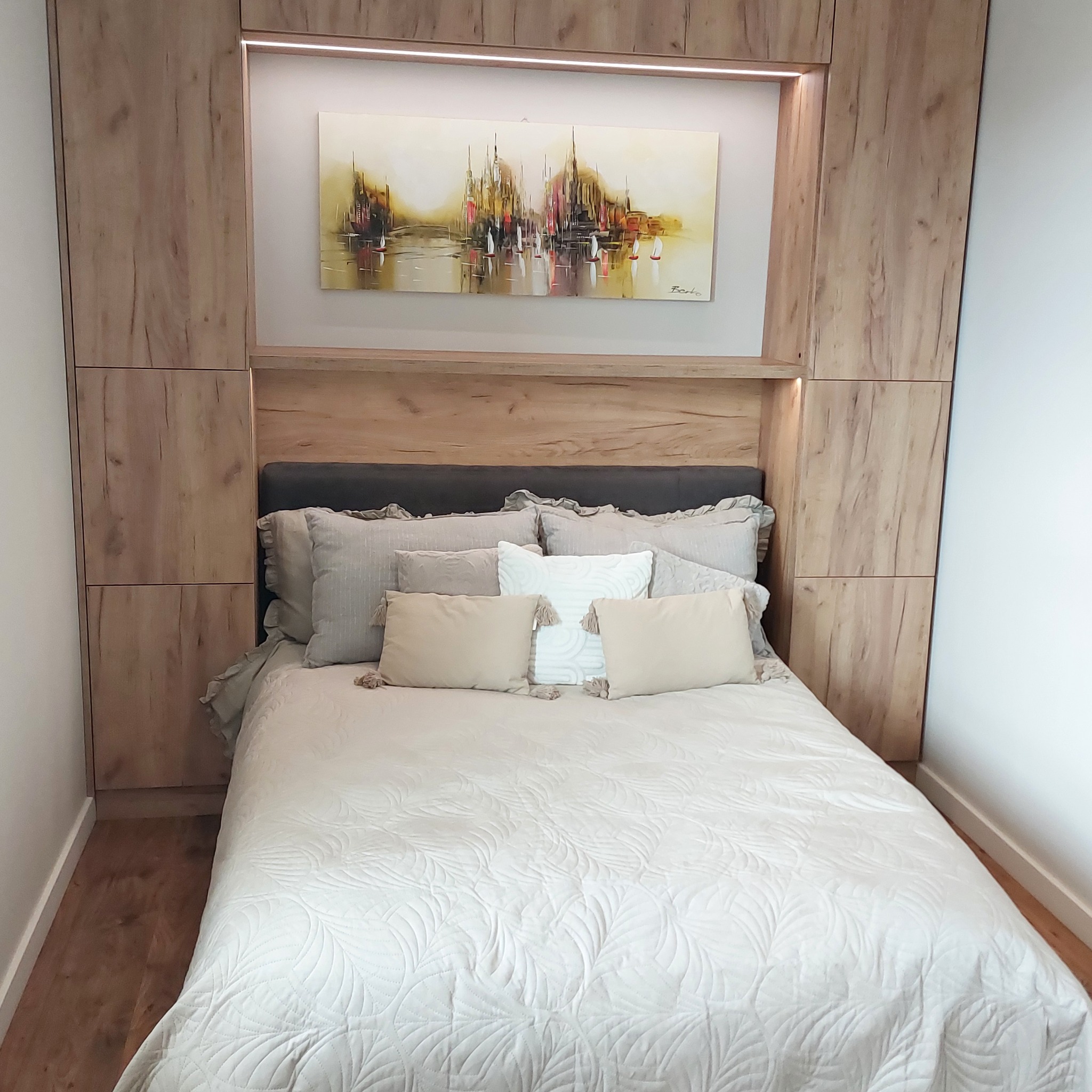 Drewniana zabudowa łóżka z szafkami i półkami, oświetlenie LED, obraz z motywem żaglówek. Jasna pościel i poduszki. Aranżacja sypialni w neutralnych barwach.