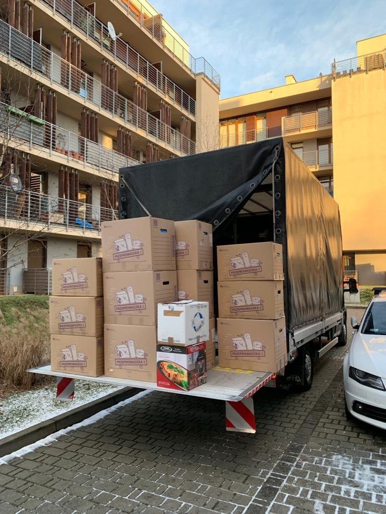 Załadunek kartonów oznaczonych napisem 'Przeprowadzka' na platformę samochodu dostawczego z czarną plandeką, na tle bloku mieszkalnego w słoneczny dzień.