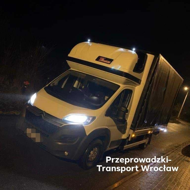 Biały bus z czarną plandeką, oświetlony reflektorami, stojący na mokrej ulicy nocą.