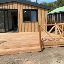 Nowoczesny taras z jasnego drewna z balustradą w trakcie montażu, przylegający do budynku z drewnianą elewacją i czarnymi ramami okiennymi.