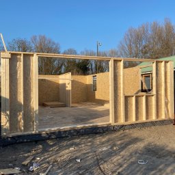 Mpower Remonty - Wczesny etap budowy drewnianej altany lub domku letniskowego: surowe ściany z płyty OSB na drewnianym szkielecie, widoczne elementy konstrukcyjne i brak wykończenia zewnętrznego.