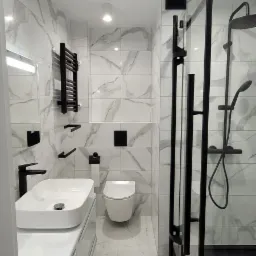 Nowoczesna łazienka z białymi marmurkowymi płytkami, czarną armaturą i kabiną prysznicową typu walk-in z czarnym brodzikiem, oświetlona punktowymi lampami sufitowymi.