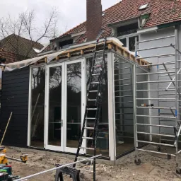 Budowa przeszklonej konstrukcji przy domu jednorodzinnym z wykorzystaniem rusztowania i drabiny, widoczne narzędzia i materiały budowlane.