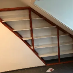 Zabudowa wnęki na poddaszu z drewnianym regałem z białymi półkami, widoczne niedoskonałości wykończenia przy ścianie.