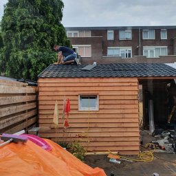 Mpower Remonty - Mężczyzna w trakcie układania czarnej dachówki na drewnianej konstrukcji budynku gospodarczego z oknem, widok na posesję i blok mieszkalny w tle.