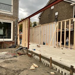 Mpower Remonty - Budowa drewnianego tarasu: szkielet konstrukcji z widoczną stalową belką, fundamentem i dwoma pracownikami w trakcie montażu na tle budynków mieszkalnych.