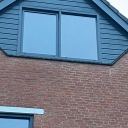 Mpower Remonty - Fragment ściany domu z czerwonej cegły z oknem w szczycie, obłożonym ciemnoszarą drewnianą boazerią.