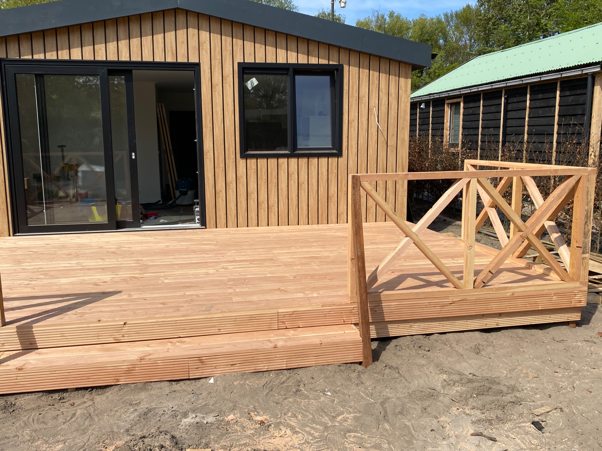 Nowoczesny taras z jasnego drewna z balustradą w trakcie montażu, przylegający do budynku z drewnianą elewacją i czarnymi ramami okiennymi.