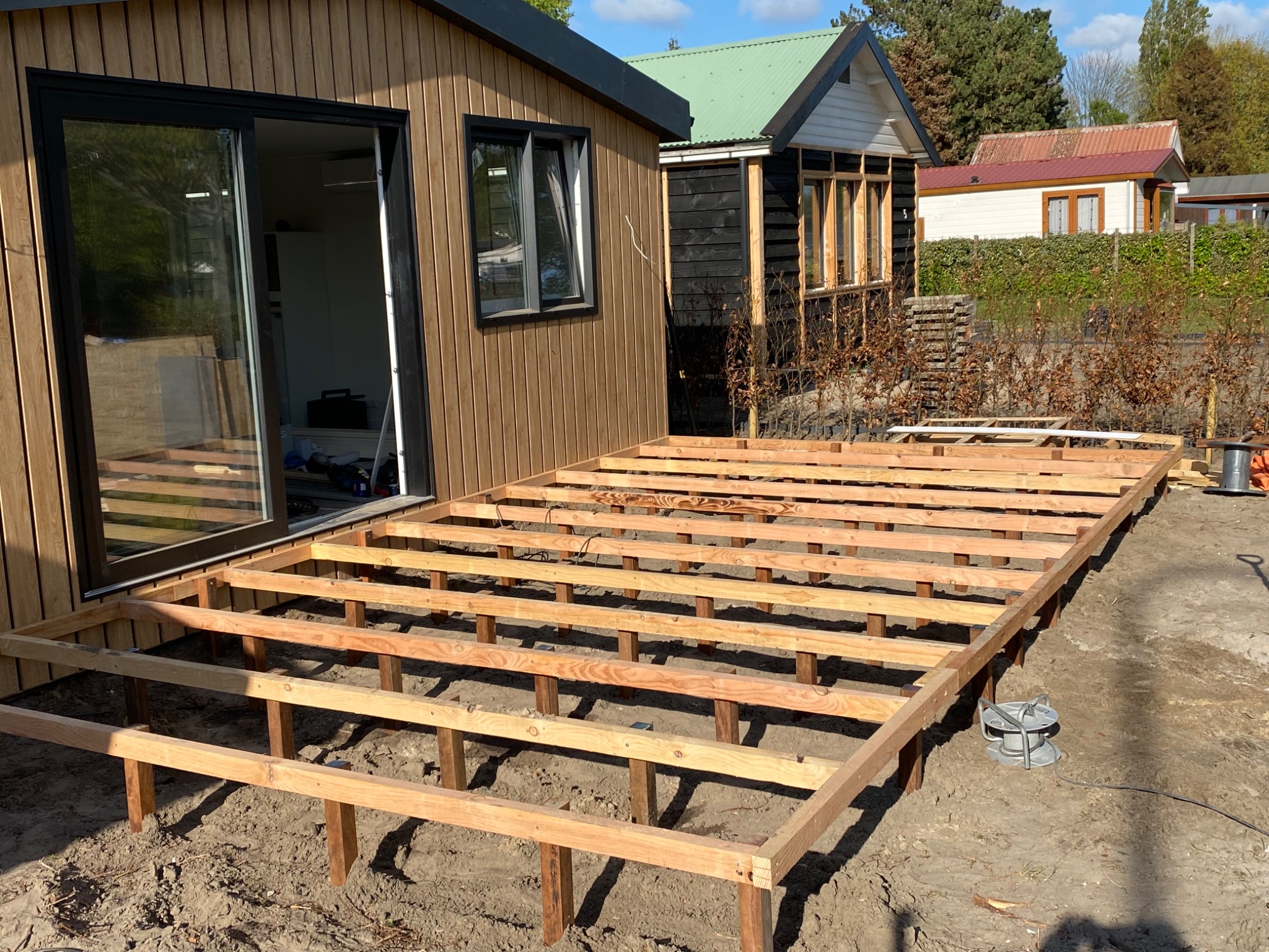 Konstrukcja drewnianego tarasu w trakcie budowy w Wałbrzychu, widoczne legary i słupy nośne na gruncie, w tle budynek z przesuwnymi drzwiami i inne zabudowania.