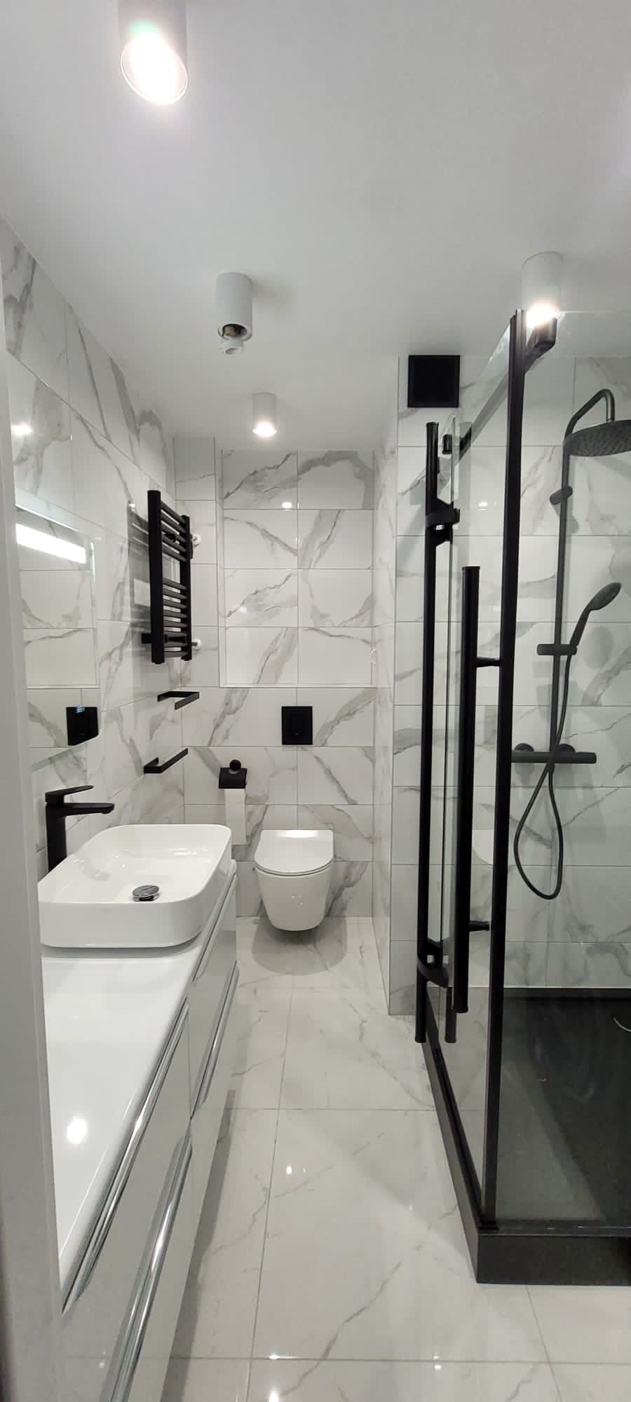 Nowoczesna łazienka z białymi marmurkowymi płytkami, czarną armaturą i kabiną prysznicową typu walk-in z czarnym brodzikiem, oświetlona punktowymi lampami sufitowymi.