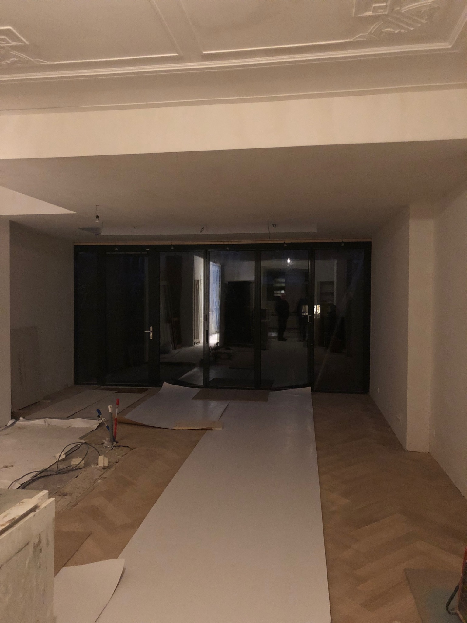 Wnętrze w trakcie remontu z widoczną podłogą ułożoną w jodełkę, zabezpieczoną częściowo białą folią, oraz zamontowanymi szklanymi drzwiami przesuwnymi z czarnymi ramami; widoczne przewody i narzędzia.