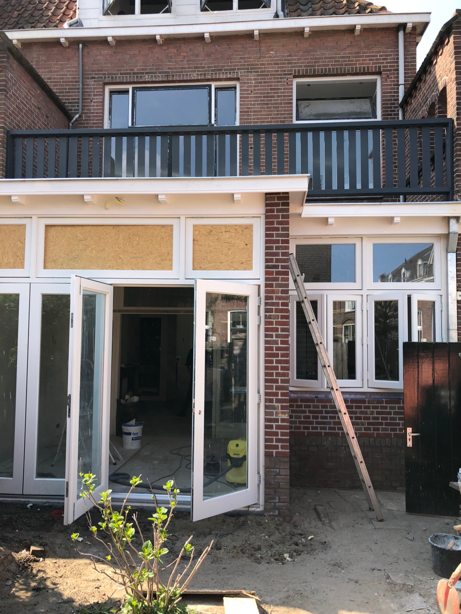 Rozbudowa domu z cegły: widoczne otwarte, przeszklone drzwi balkonowe, drewniana drabina oparta o ścianę, częściowo zabite okna płytami OSB, balkon z czarną balustradą na piętrze.