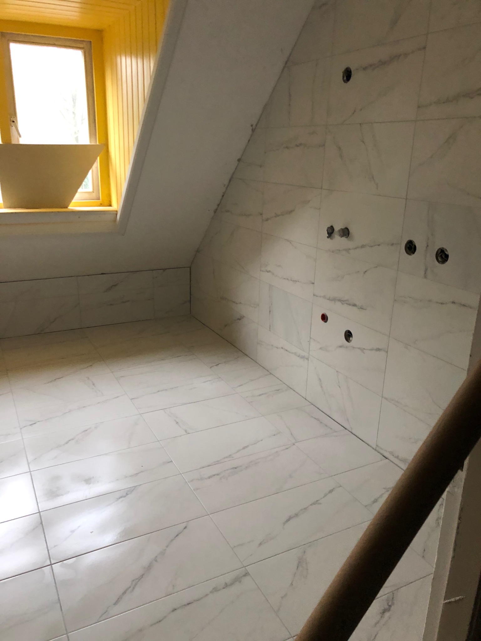Łazienka w trakcie remontu: białe marmurowe płytki na podłodze i ścianach, widoczne wyprowadzenia instalacji wodnej, żółta rama okienna z nietypową donicą.