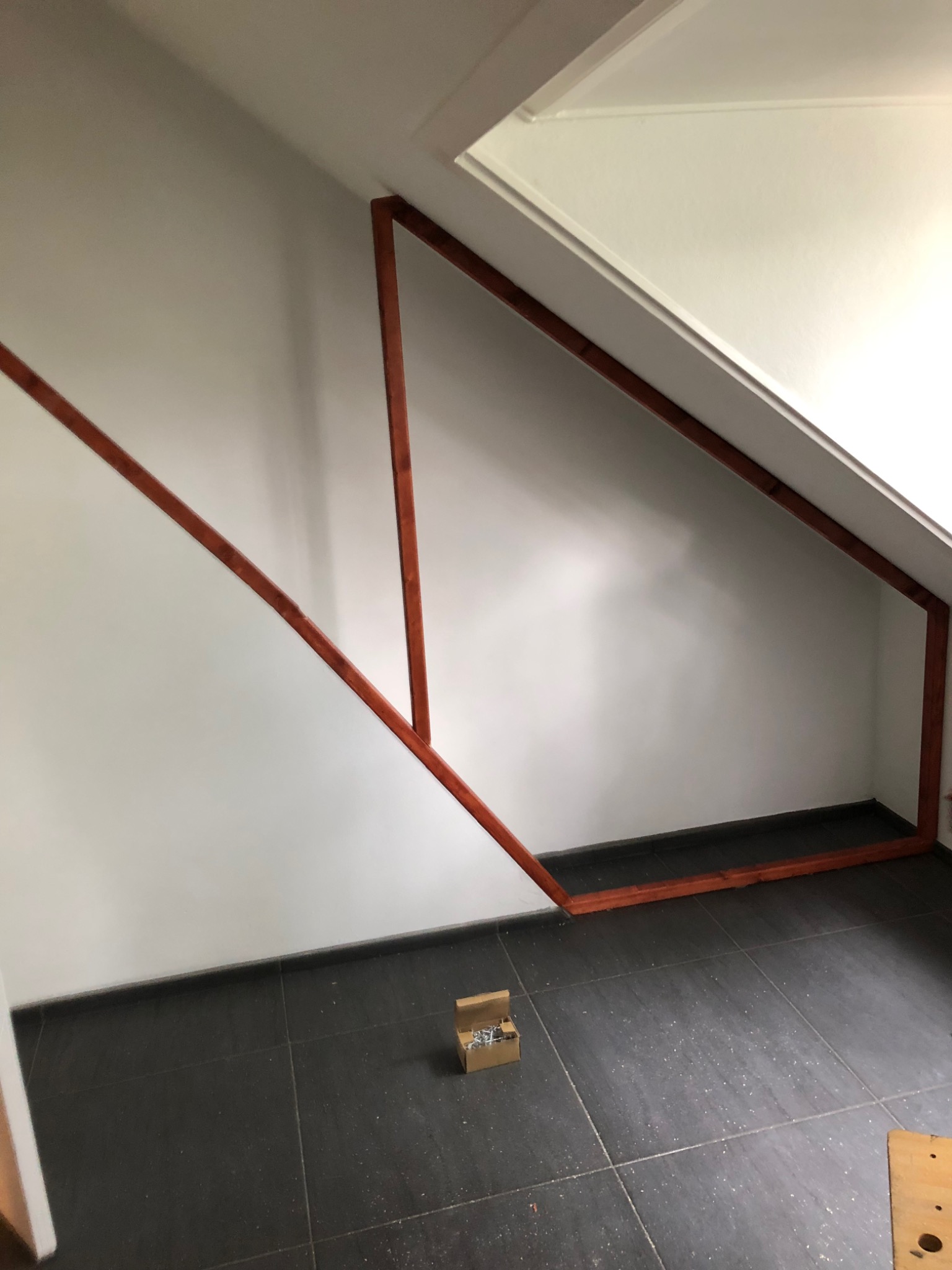 Drewniana konstrukcja w trakcie montażu na poddaszu ze skosem, widoczna skrzynka ze śrubami na ciemnej podłodze z płytek.