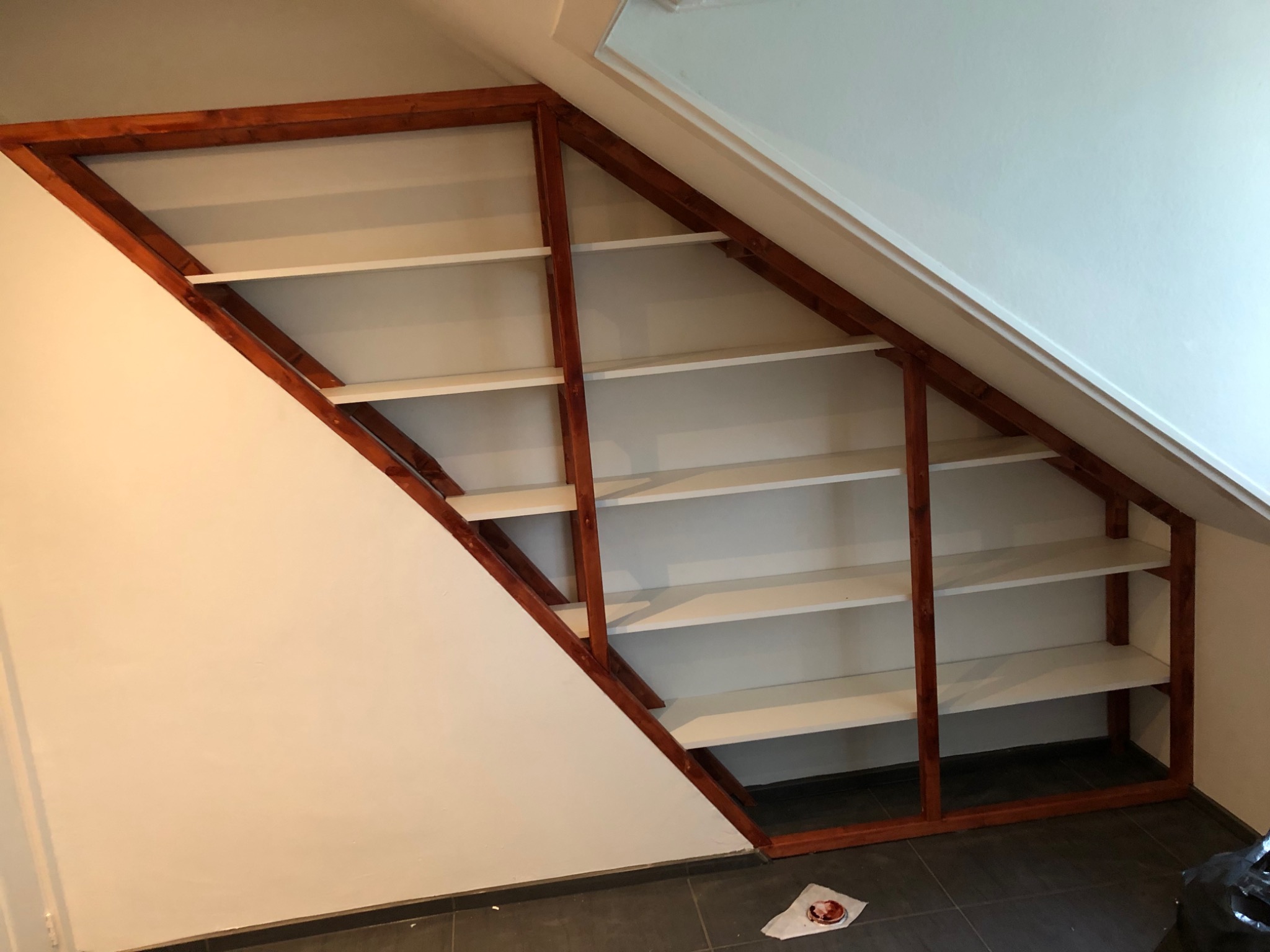 Zabudowa wnęki na poddaszu z drewnianym regałem z białymi półkami, widoczne niedoskonałości wykończenia przy ścianie.