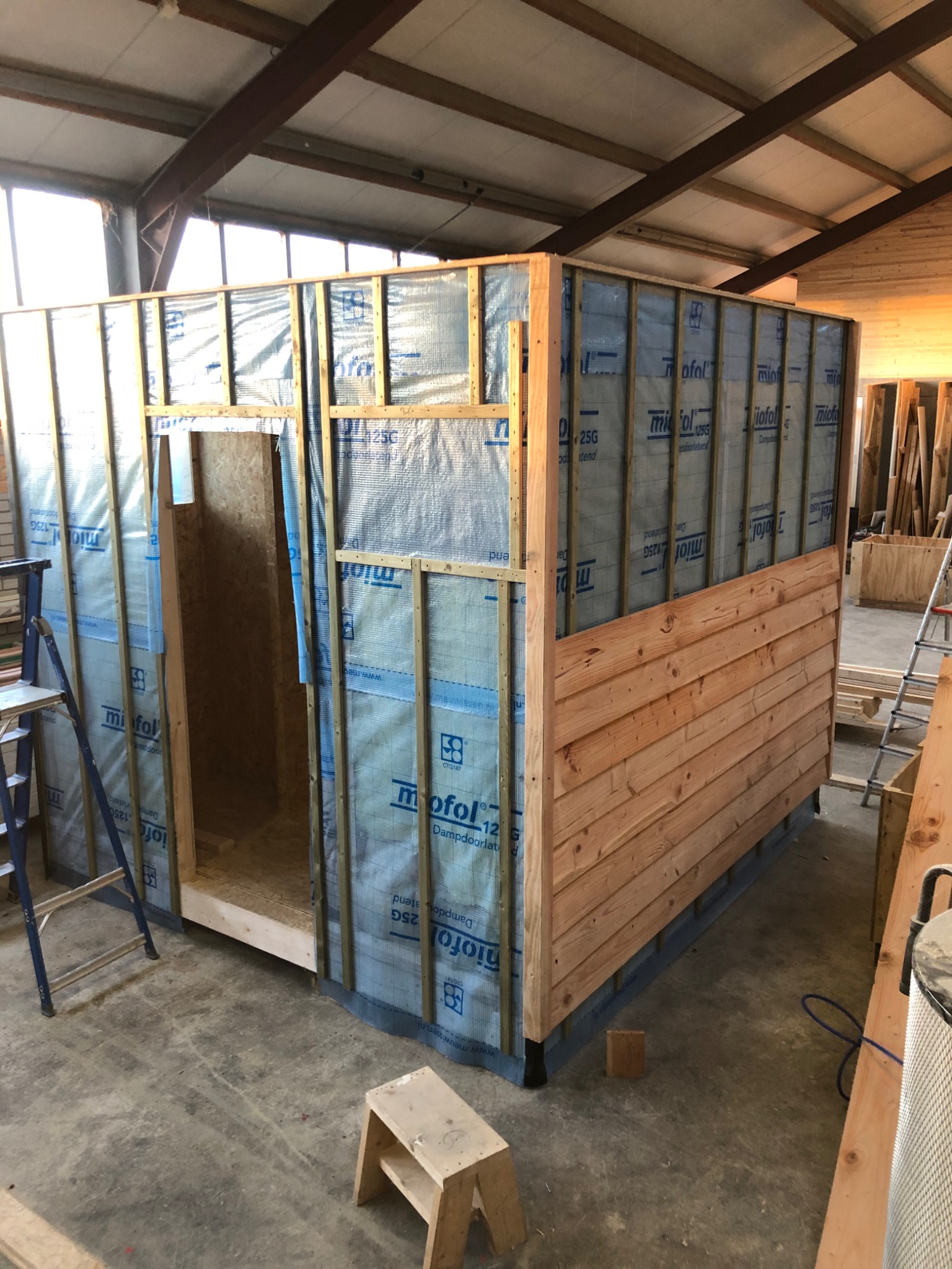 Wnętrze hali z widoczną konstrukcją ścianki działowej w trakcie budowy, częściowo obłożoną drewnianą boazerią, okrytą folią paroizolacyjną, z widocznym wejściem, drabina i stołek w tle.