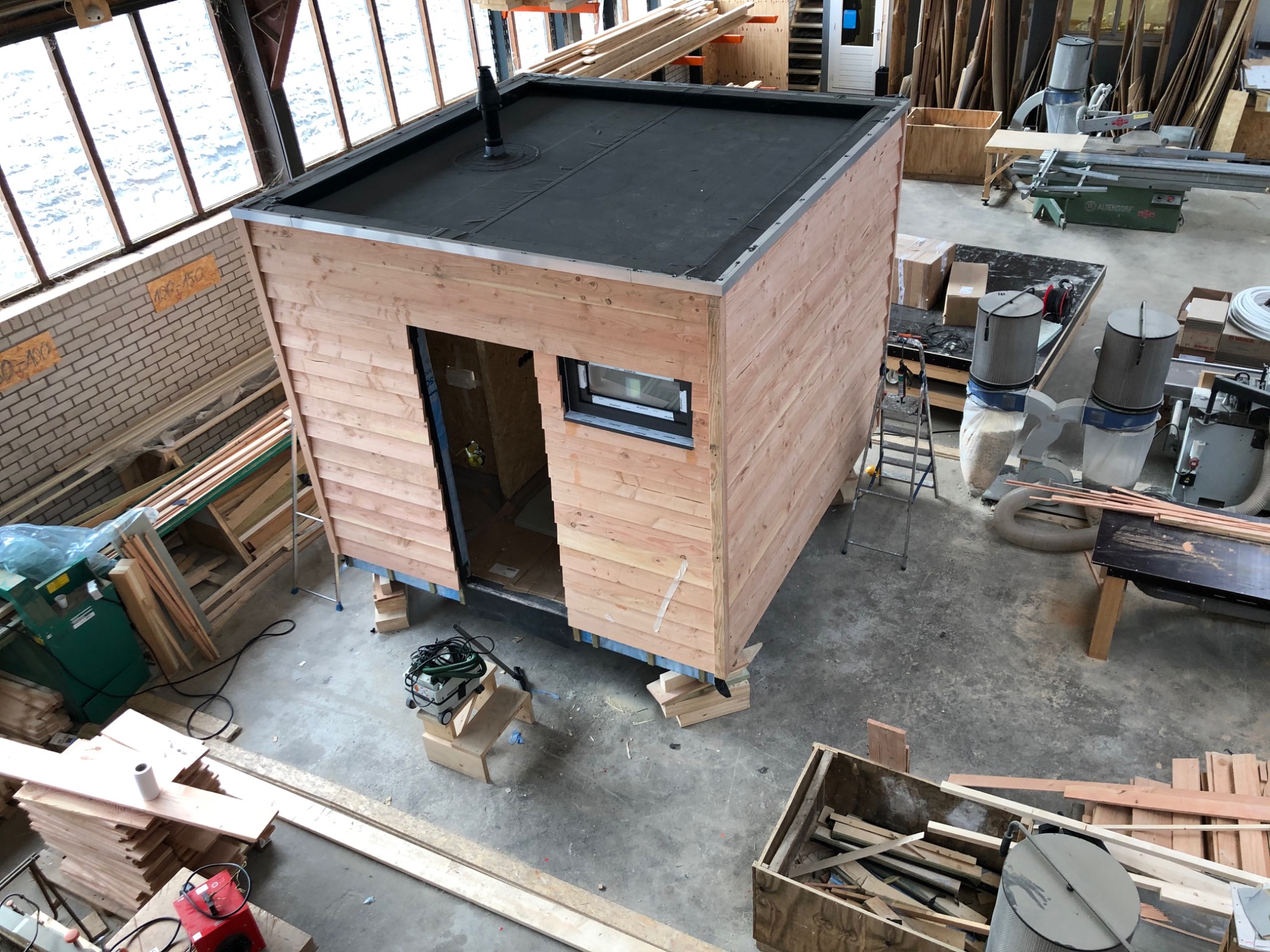 Wnętrze warsztatu z widokiem z góry na częściowo zmontowaną, drewnianą altanę z płaskim dachem pokrytym papą, umieszczoną na drewnianych podporach, z widocznym oknem i otwartymi drzwiami.