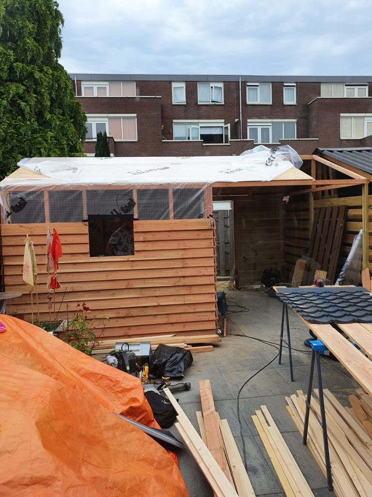 W trakcie budowy drewniana konstrukcja w ogrodzie, widoczne deski, narzędzia i osoba w środku, dach zabezpieczony folią, w tle budynek mieszkalny.
