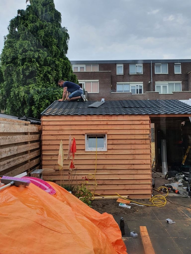 Mężczyzna w trakcie układania czarnej dachówki na drewnianej konstrukcji budynku gospodarczego z oknem, widok na posesję i blok mieszkalny w tle.