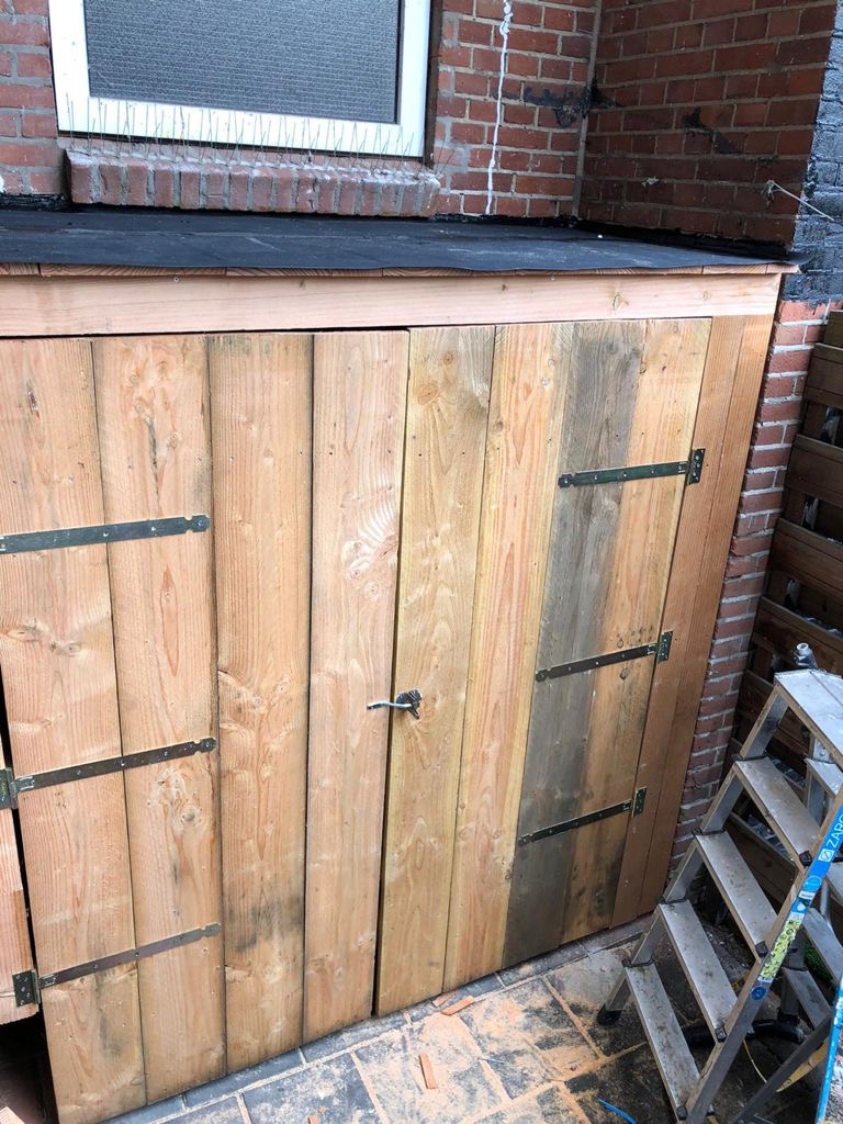 Drewniana zabudowa z dwuskrzydłowymi drzwiami, przymocowana do ściany z cegły, z widocznym oknem zabezpieczonym kolcami i drabinką obok.