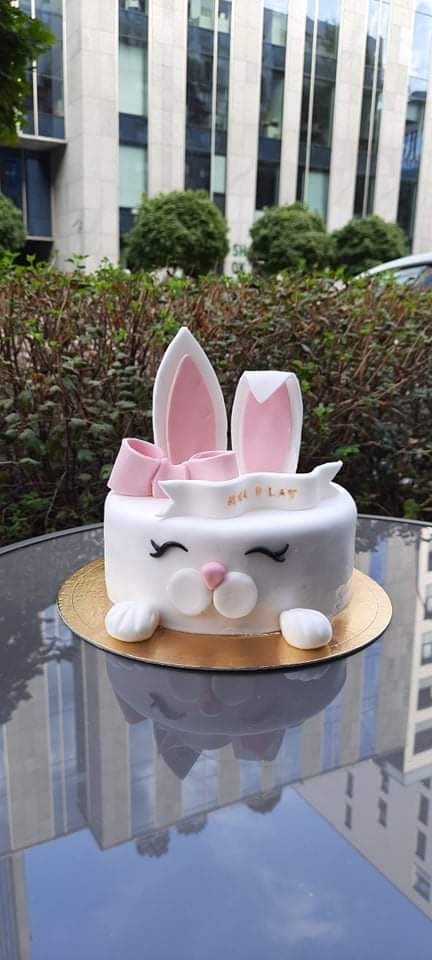 Tort w kształcie królika z białym lukrem, różowymi uszami i kokardką, na złotym podkładzie, z napisem 'Play', sfotografowany na tle budynku biurowego i zieleni.