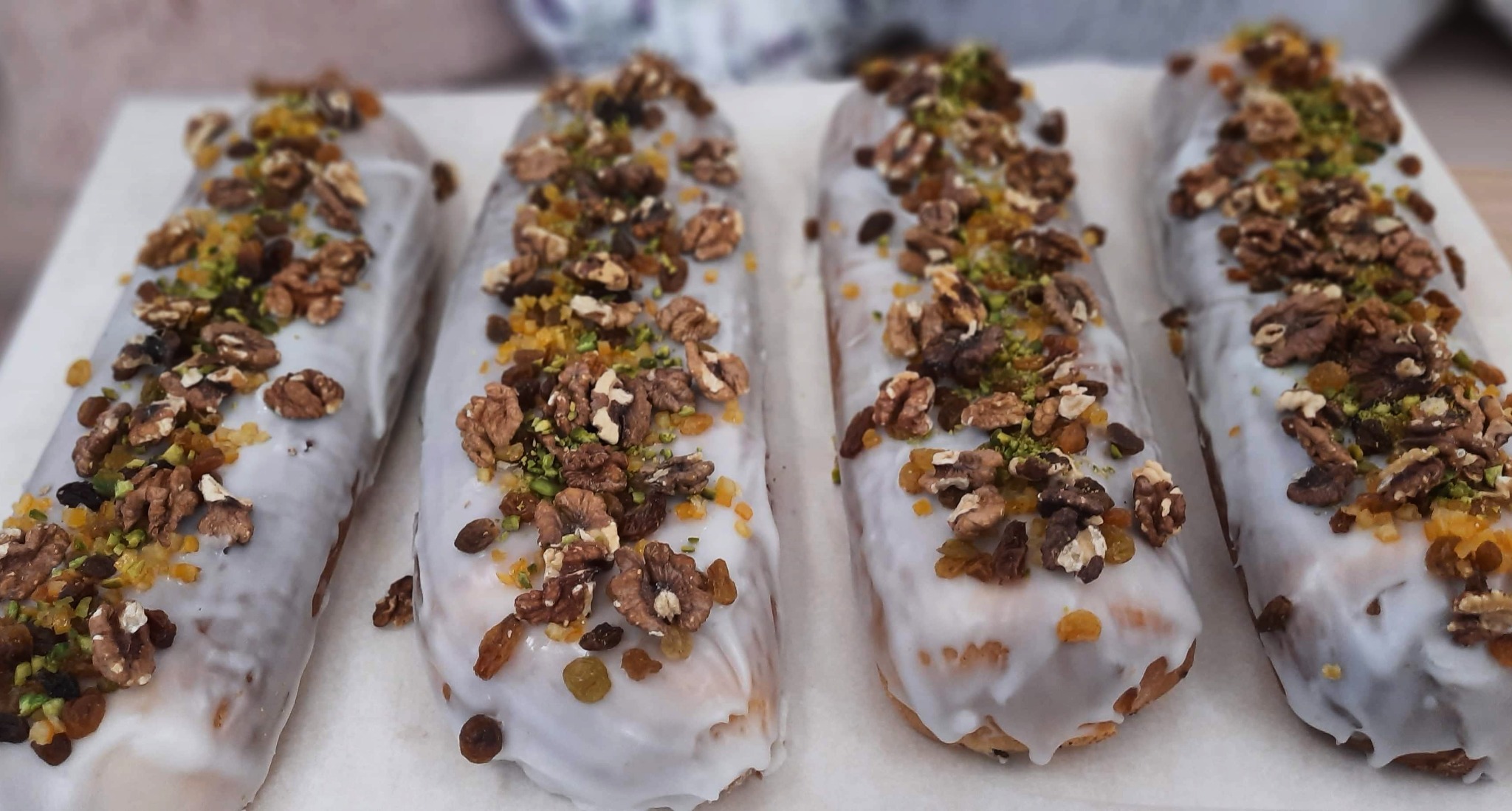 Cztery keksowe babki polane białym lukrem i posypane orzechami włoskimi, pistacjami i kandyzowaną skórką pomarańczową, ułożone na białym papierze.