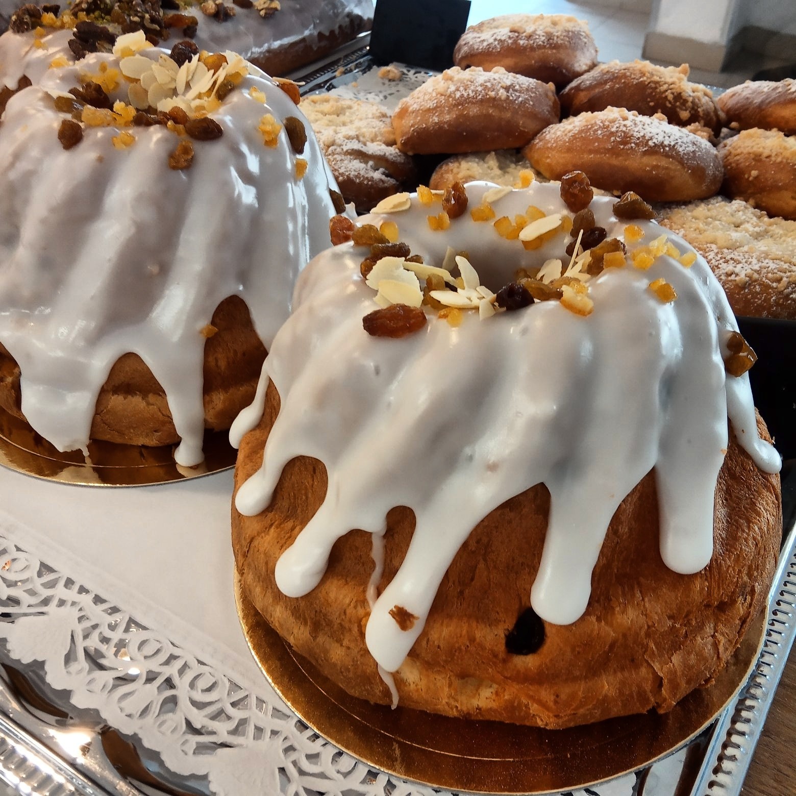 Dwie babki wielkanocne z białym lukrem, posypane rodzynkami, skórką pomarańczową i płatkami migdałów, na srebrnej tacy z koronkową serwetką, w tle bułeczki posypane kruszonką.