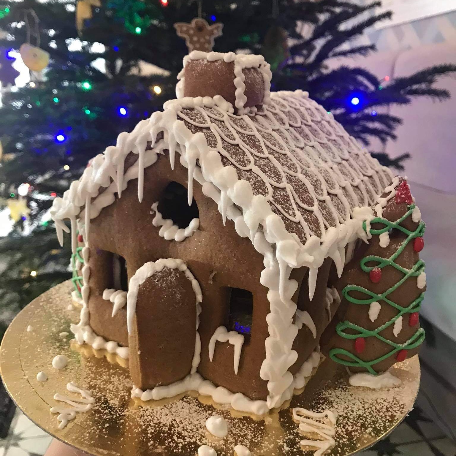 Piernikowa chatka z lukrowanymi detalami i choinką z piernika, na złotym talerzu, w tle choinka świąteczna z kolorowymi światełkami.