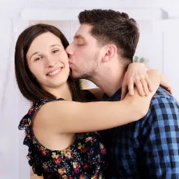 Uśmiechnięta kobieta w kwiecistej sukience obejmowana przez mężczyznę w niebieskiej koszuli w kratę, który całuje ją w policzek na tle białego studia.