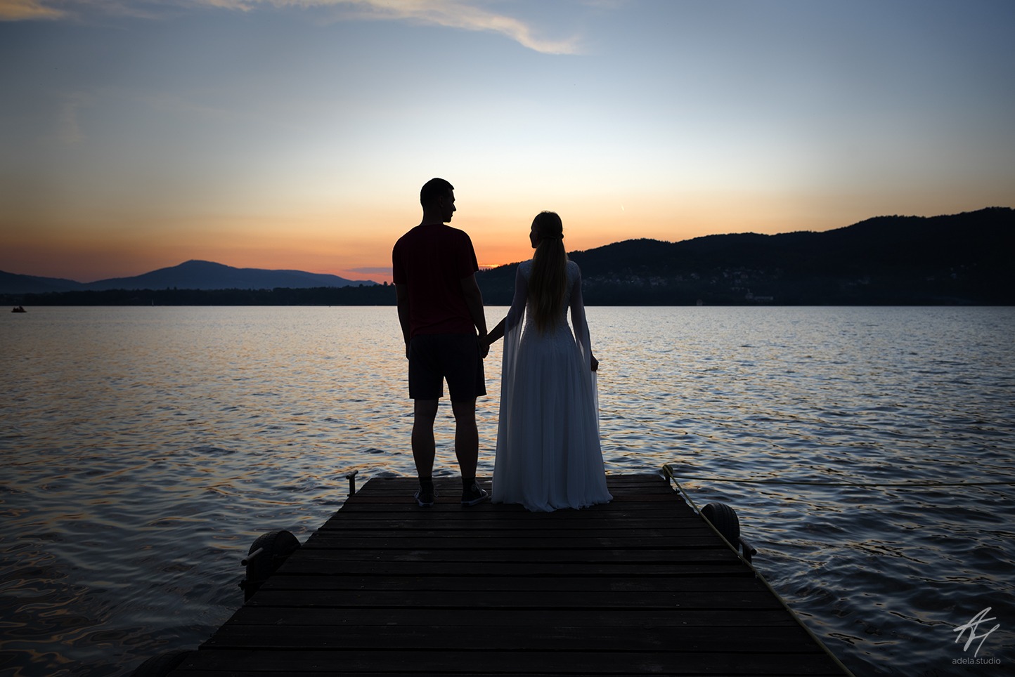 Para młoda, stojąca tyłem na drewnianym pomoście, trzyma się za ręce, obserwując zachód słońca nad jeziorem i zalesionymi wzgórzami w oddali.