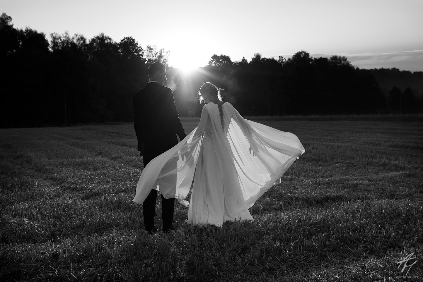 Czarno-białe zdjęcie pary młodej, stojącej tyłem na polu o zachodzie słońca. Panna młoda w zwiewnej sukni z peleryną, delikatnie podświetlona promieniami słońca.