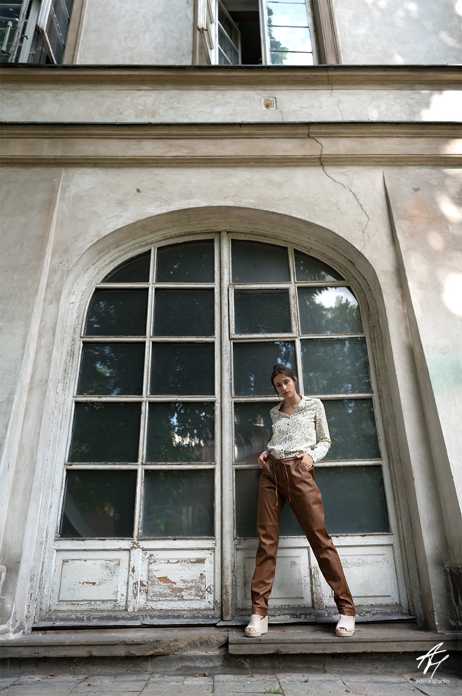 Kobieta w koszuli we wzory i skórzanych spodniach pozuje na tle dużych, przeszklonych drzwi z łukowym nadprożem i widocznymi śladami użytkowania.