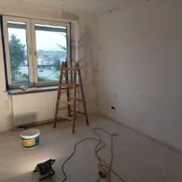 Pokój w trakcie szpachlowania ścian, widoczny drewniany drabina, wiadro z masą szpachlową, lampa halogenowa na podłodze, ściany z widocznymi miejscami po szpachlowaniu, okno z widokiem na zabudowania.