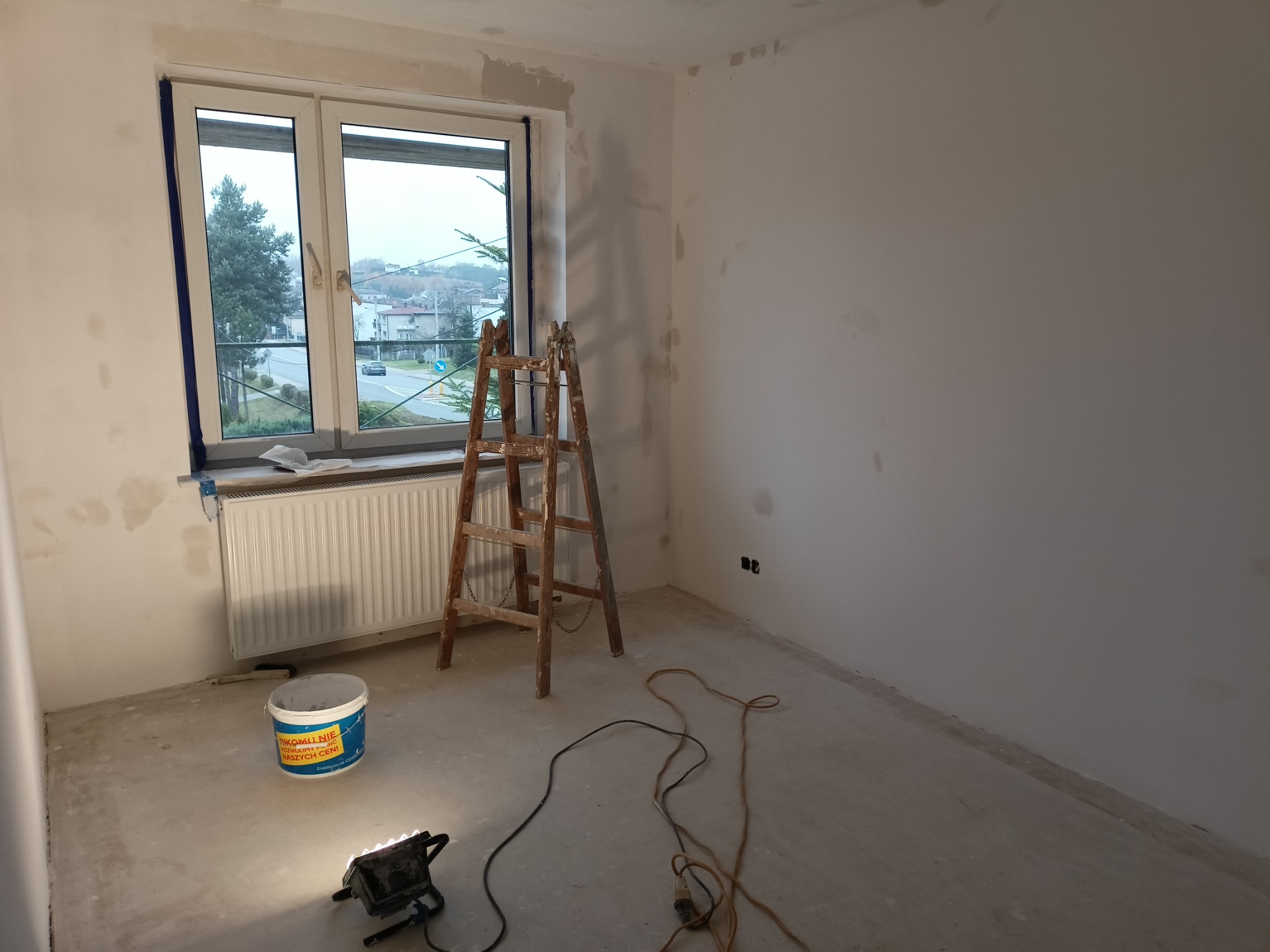 Pokój w trakcie szpachlowania ścian, widoczny drewniany drabina, wiadro z masą szpachlową, lampa halogenowa na podłodze, ściany z widocznymi miejscami po szpachlowaniu, okno z widokiem na zabudowania.