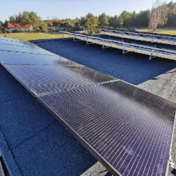 Panele słoneczne w trakcie montażu na płaskim dachu pokrytym papą, widoczne metalowe konstrukcje wsporcze i elementy mocujące, w tle zabudowa jednorodzinna i drzewa.