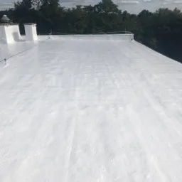 Biała, płaska powierzchnia dachu pokryta jednolitą warstwą izolacji natryskowej, widoczne kominy i system odwadniający, w tle zieleń drzew i błękitne niebo z chmurami.