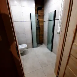 Wykończona łazienka z kabiną prysznicową typu walk-in z czarnymi ramami, białą ceramiką i grzejnikiem drabinkowym widziana zza uchylonych drewnianych drzwi.