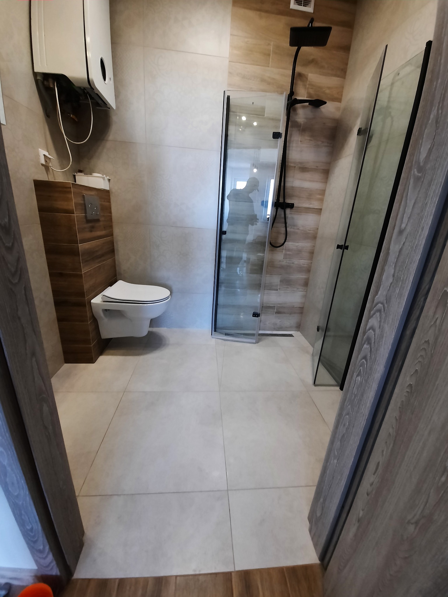 Nowoczesna łazienka po remoncie z kabiną prysznicową typu walk-in z czarną armaturą, podwieszaną toaletą i grzejnikiem gazowym.