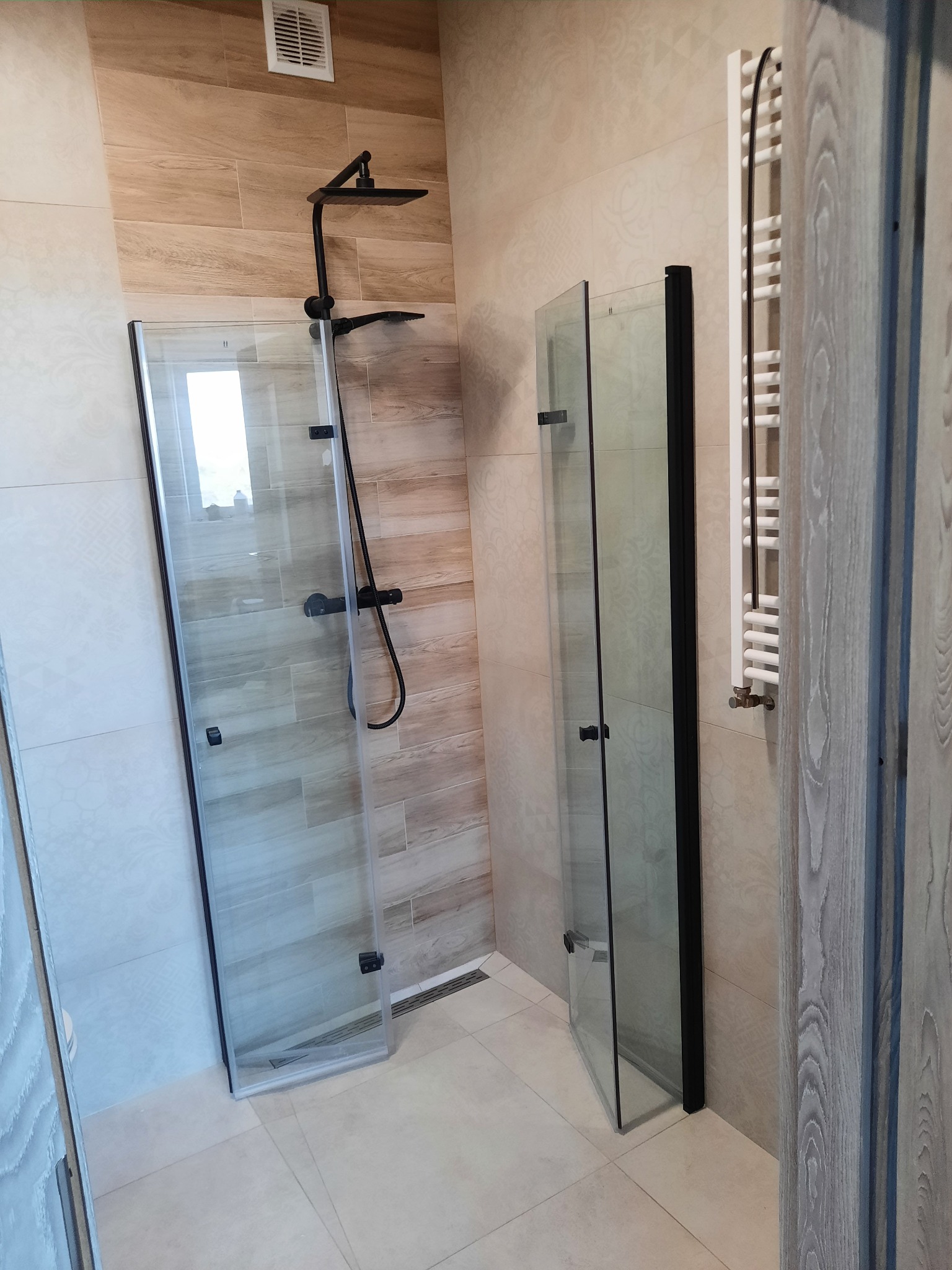 Nowoczesna kabina prysznicowa walk-in z czarnymi profilami i deszczownicą, płytki imitujące drewno na ścianie, grzejnik drabinkowy w łazience z jasnymi płytkami.