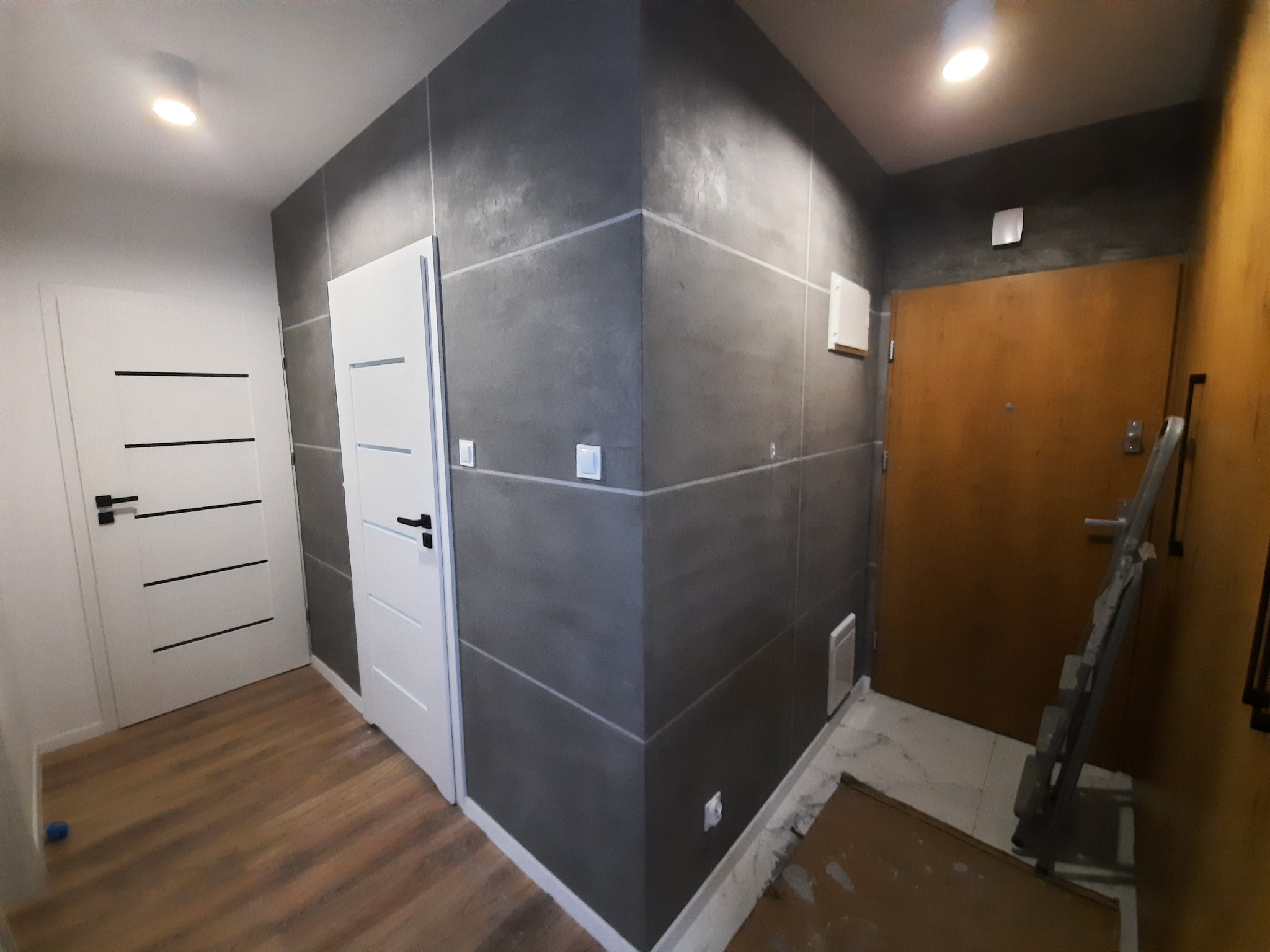 Nowoczesny przedpokój po remoncie: ściany pokryte dekoracyjną szarą strukturą, białe drzwi z poziomymi czarnymi paskami, drewnopodobna podłoga, widoczna drabina malarska przy wejściowych drzwiach.