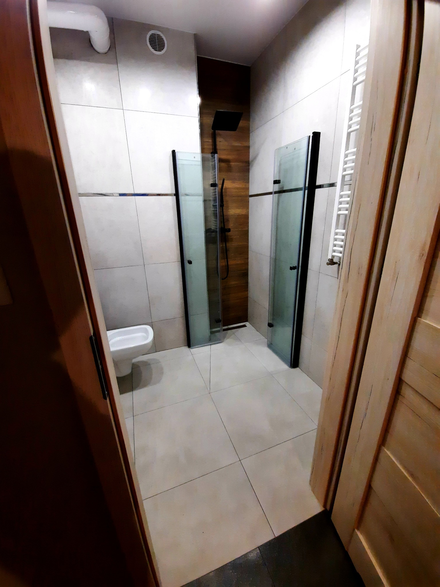 Wykończona łazienka z kabiną prysznicową typu walk-in z czarnymi ramami, białą ceramiką i grzejnikiem drabinkowym widziana zza uchylonych drewnianych drzwi.