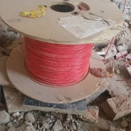 Szpula z czerwonym kablem elektrycznym na budowie, otoczona gruzem i fragmentami cegieł, przy ścianie z surowej cegły.