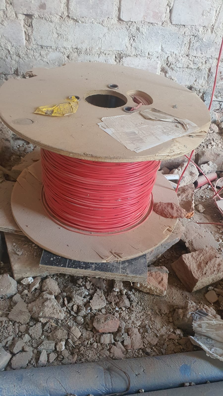 Szpula z czerwonym kablem elektrycznym na budowie, otoczona gruzem i fragmentami cegieł, przy ścianie z surowej cegły.