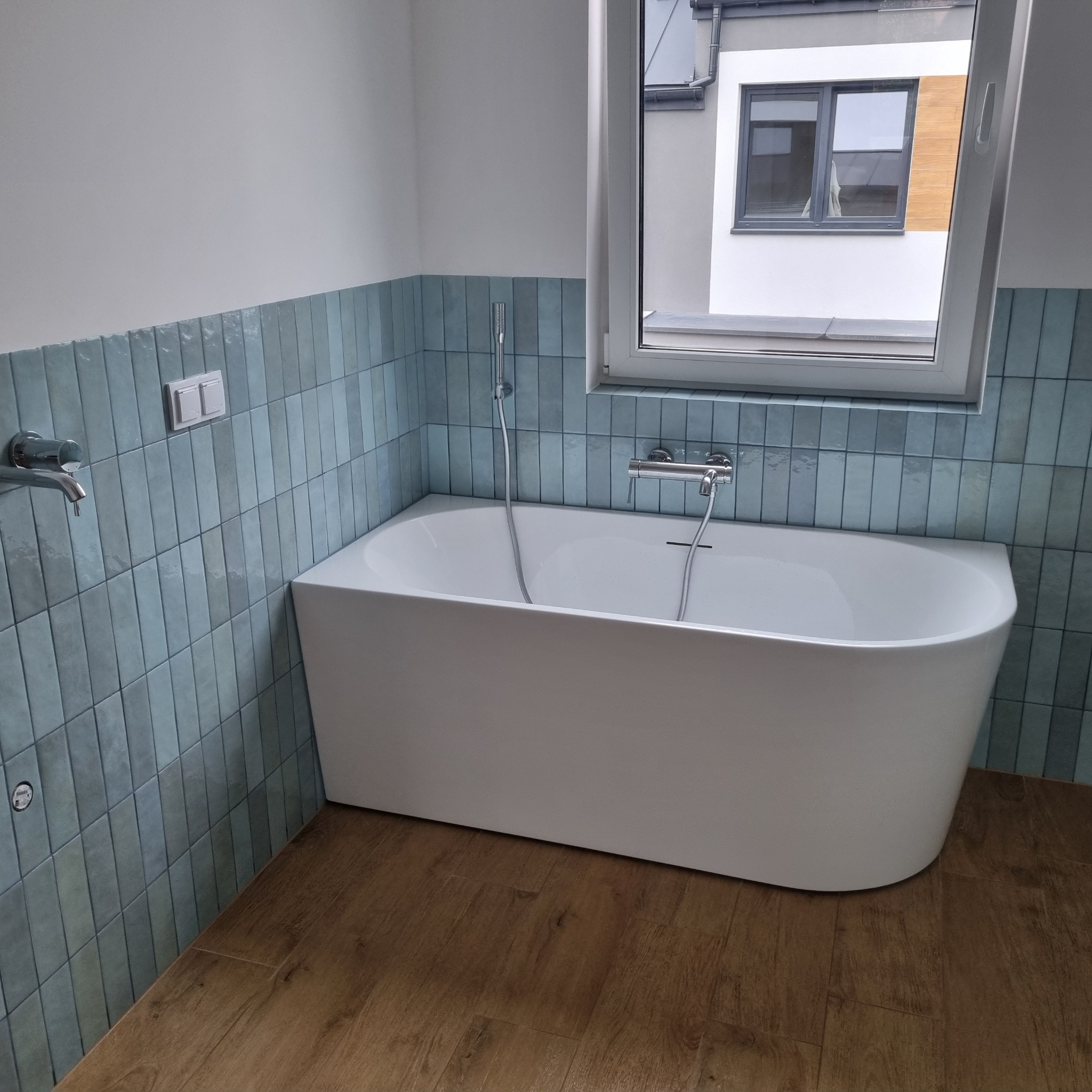 Biała wanna wolnostojąca na tle ściany z niebieskich, pionowo ułożonych płytek, z widocznym oknem i drewnopodobną podłogą.