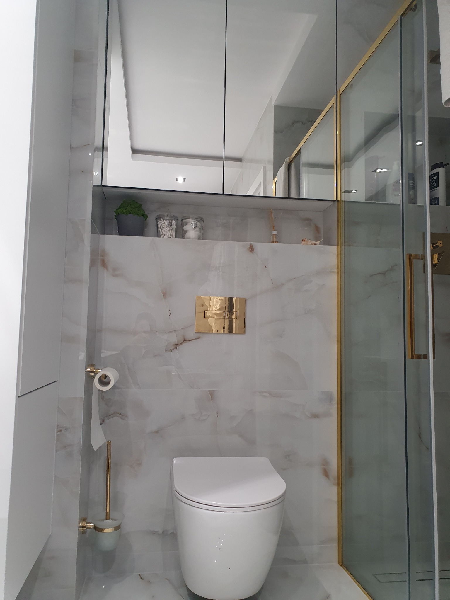 Nowoczesna łazienka z białym sedesem, marmurowymi płytkami, złotymi akcentami na armaturze i kabinie prysznicowej, oraz szklanymi szafkami z rośliną i pojemnikami na akcesoria.