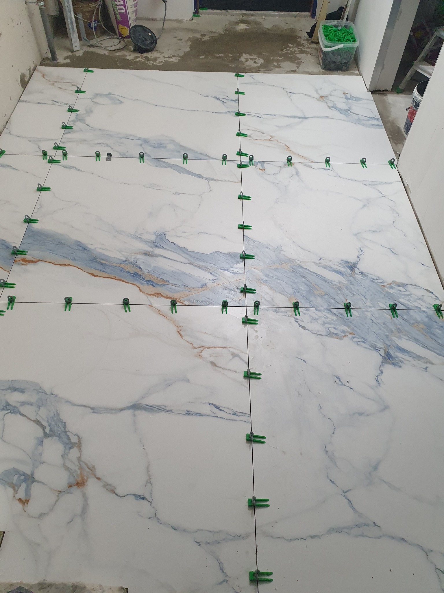 Układanie dużych, białych płytek imitujących marmur z niebieskimi i brązowymi żyłami na podłodze, z użyciem zielonych klipsów poziomujących i czarnych sznurków dystansowych, widoczny etap prac.